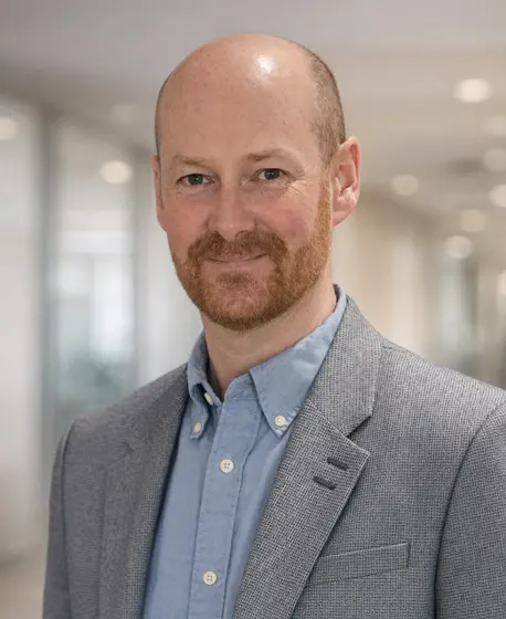 Professional headshot of a Mark McKnight in a grey blazer and blue shirt, smiling in a modern office corridor.